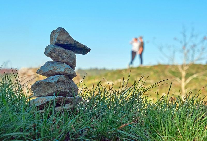 Aufgeschichteter Steinturm im Gras, im Hintergrund ein Paar auf einem Hügel unter blauem Himmel